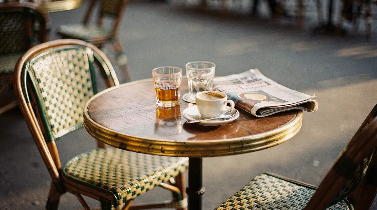 Table de terrasse de café ensoleillée : deux verres, une tasse d'espresso et un journal plié, deux chaises en cannage