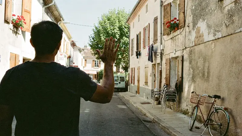Homme cadré à mi-corps, de dos, la main levée en salut vers une rue ensoleillée où il n'y a personne