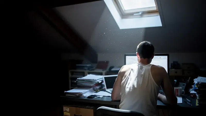 Homme de dos en marcel à son bureau mansardé, le rai de lumière d'une lucarne tombant sur son épaule pâle