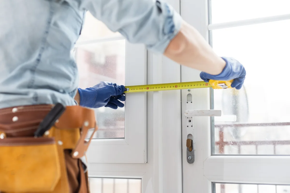 Artisan du bâtiment mesurant un cadre de fenêtre avec un mètre ruban, ceinture à outils
